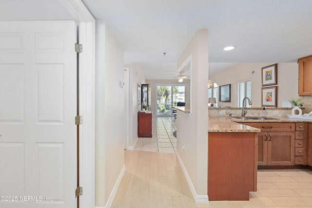 kitchen with baseboards, light stone countertops, a peninsula, brown cabinetry, and a sink