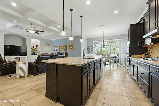 kitchen with light tile patterned floors, a center island with sink, arched walkways, a sink, and beamed ceiling