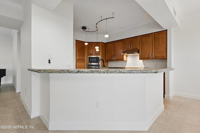 kitchen with under cabinet range hood, stainless steel microwave, brown cabinets, and light stone counters