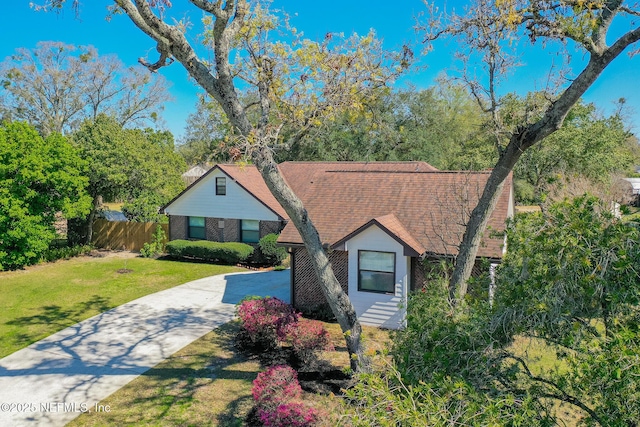 view of front facade featuring brick siding, fence, roof with shingles, a front yard, and driveway