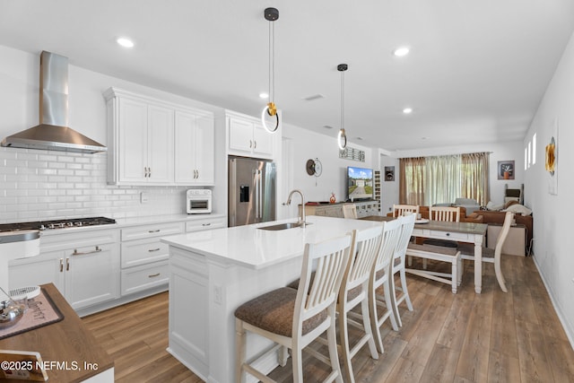 kitchen with light wood-type flooring, stainless steel refrigerator with ice dispenser, a sink, tasteful backsplash, and wall chimney exhaust hood