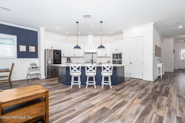 kitchen featuring light countertops, appliances with stainless steel finishes, white cabinetry, an island with sink, and custom range hood