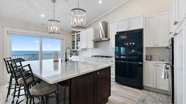 kitchen with wall chimney range hood, an island with sink, freestanding refrigerator, gas cooktop, and a sink