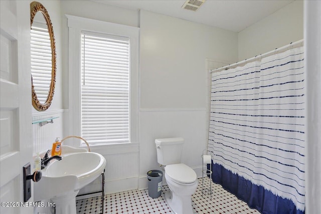 full bath featuring visible vents, toilet, curtained shower, wainscoting, and a decorative wall