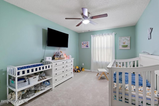 bedroom with baseboards, ceiling fan, a nursery area, a textured ceiling, and carpet flooring