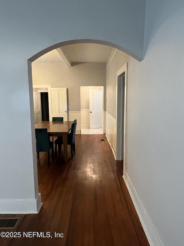 hallway with a wainscoted wall, visible vents, baseboards, dark wood finished floors, and arched walkways