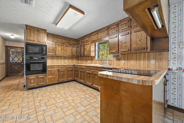 kitchen with black appliances, a sink, a peninsula, brown cabinetry, and wallpapered walls