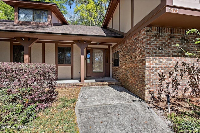 doorway to property featuring stucco siding, brick siding, covered porch, and roof with shingles