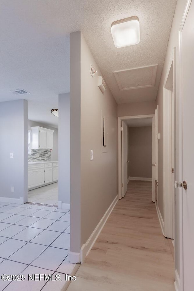 corridor featuring attic access, baseboards, light wood finished floors, and a textured ceiling