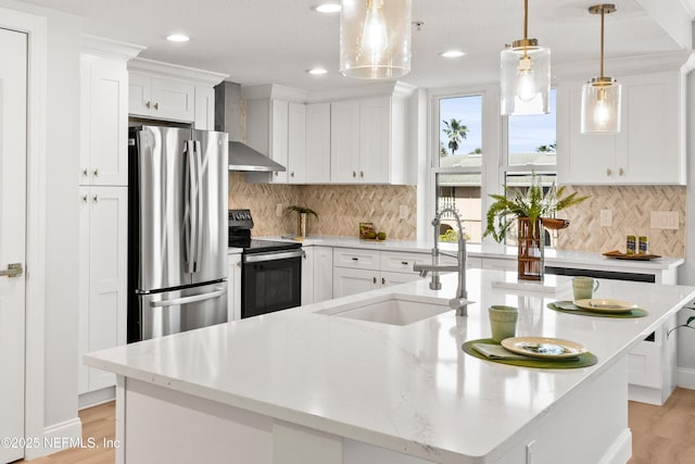 kitchen featuring an island with sink, a sink, appliances with stainless steel finishes, white cabinetry, and wall chimney range hood