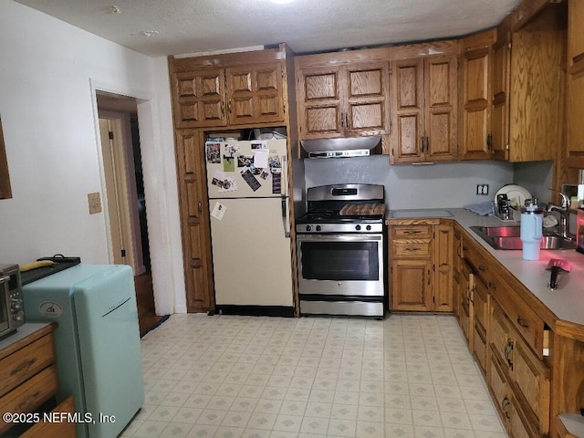 kitchen with brown cabinetry, stainless steel range with gas cooktop, freestanding refrigerator, a sink, and under cabinet range hood