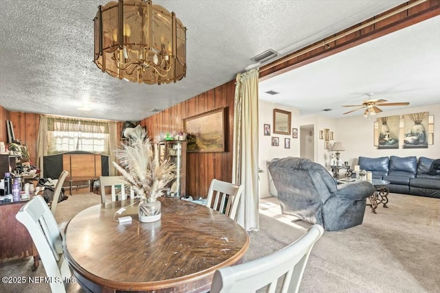 dining room featuring visible vents, ceiling fan with notable chandelier, a textured ceiling, wooden walls, and carpet