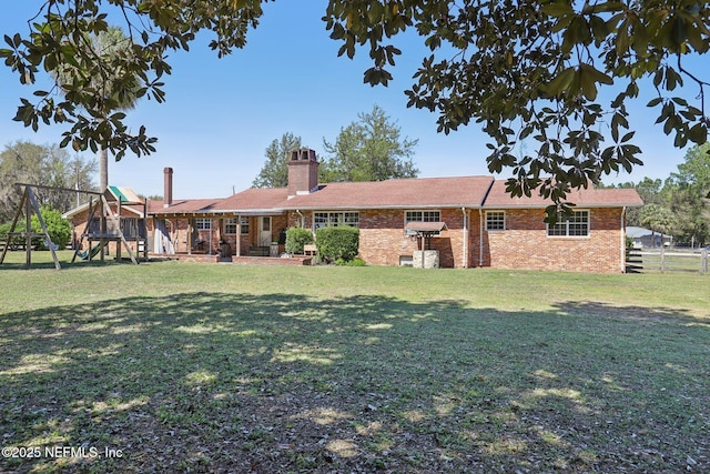 rear view of property featuring brick siding, a playground, fence, a lawn, and a chimney