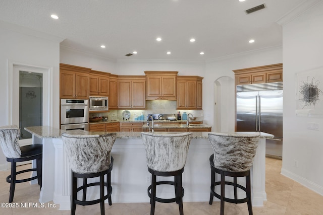 kitchen featuring visible vents, built in appliances, decorative backsplash, a large island with sink, and brown cabinetry