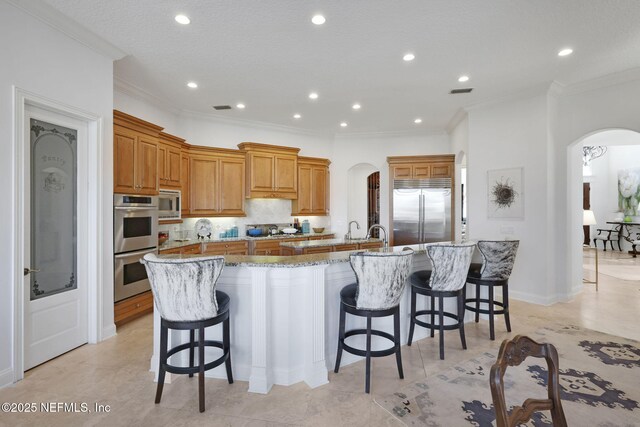 kitchen featuring light stone counters, a spacious island, arched walkways, built in appliances, and a kitchen breakfast bar