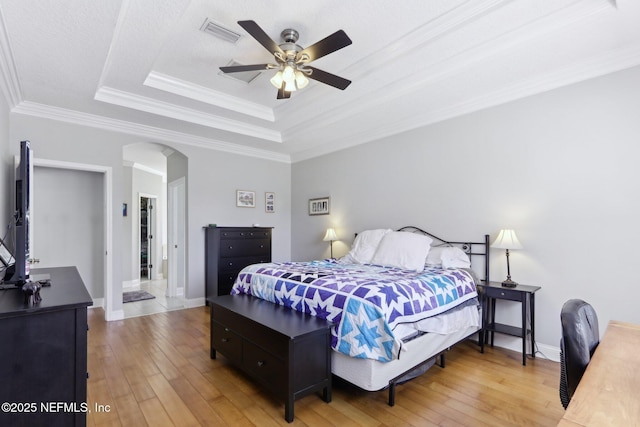 bedroom featuring visible vents, a tray ceiling, arched walkways, crown molding, and light wood-type flooring