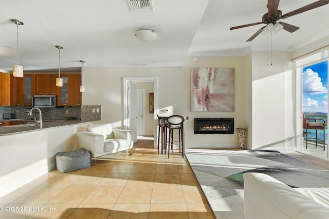 living room featuring light tile patterned floors, a ceiling fan, visible vents, a glass covered fireplace, and crown molding