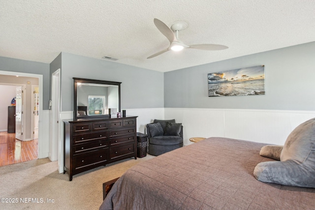 bedroom featuring visible vents, light carpet, wainscoting, a textured ceiling, and a ceiling fan