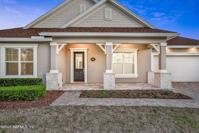 view of exterior entry with an attached garage, covered porch, a shingled roof, stucco siding, and decorative driveway