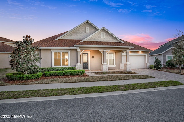 craftsman-style home with roof with shingles, an attached garage, covered porch, stucco siding, and decorative driveway