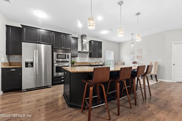 kitchen with dark cabinetry, wood finished floors, a center island with sink, stainless steel appliances, and wall chimney range hood