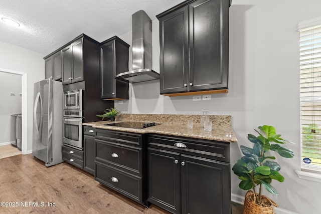 kitchen featuring light stone counters, dark cabinetry, light wood-style floors, stainless steel appliances, and wall chimney exhaust hood