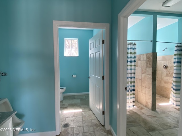 bathroom featuring vanity, baseboards, a tile shower, stone tile flooring, and toilet