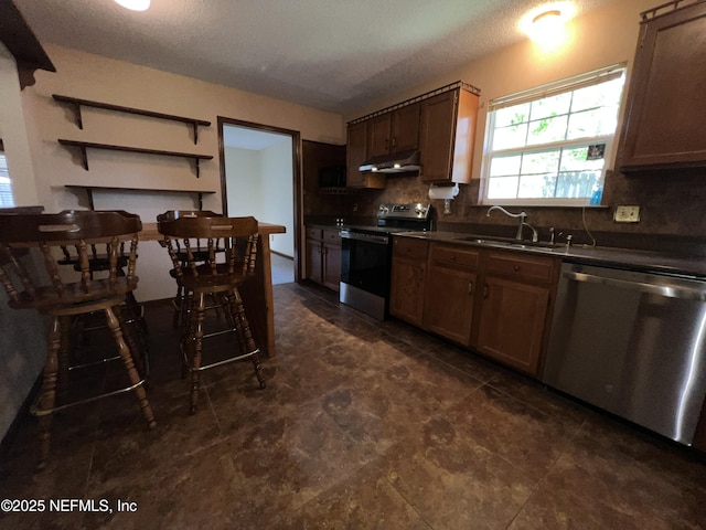 kitchen with dark countertops, under cabinet range hood, decorative backsplash, appliances with stainless steel finishes, and a sink