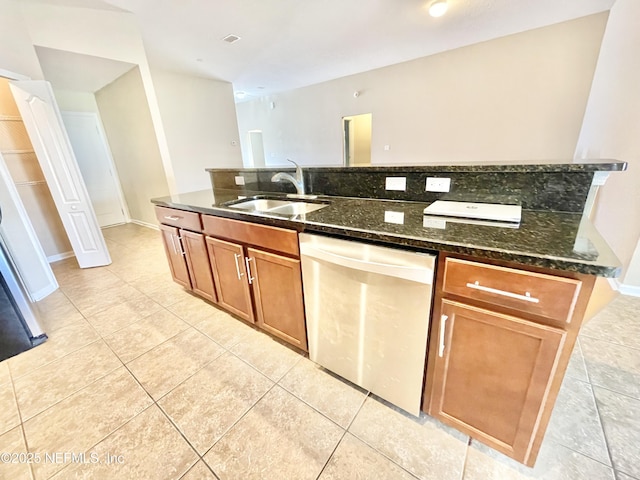 kitchen with dark stone counters, light tile patterned floors, stainless steel dishwasher, brown cabinetry, and a sink