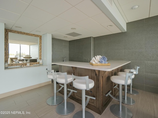 bar featuring tile walls, a community bar, baseboards, and a drop ceiling