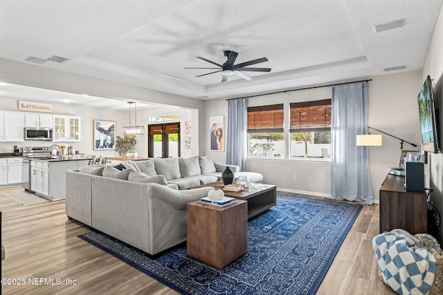 living room with ceiling fan with notable chandelier, a tray ceiling, light wood-style floors, and a textured ceiling