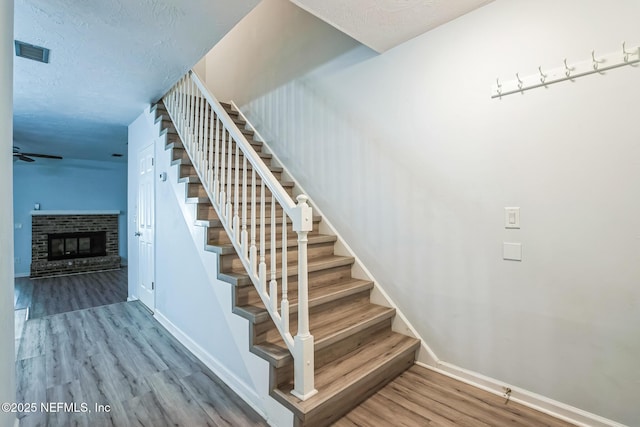 staircase featuring wood finished floors, visible vents, baseboards, a textured ceiling, and a brick fireplace