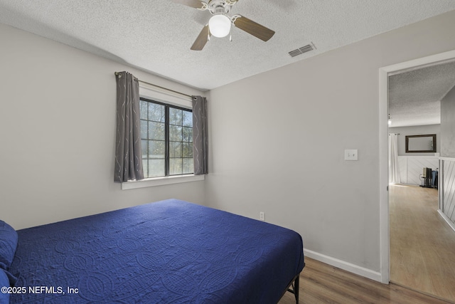 bedroom with visible vents, a textured ceiling, wood finished floors, baseboards, and ceiling fan