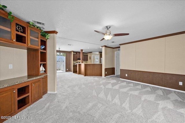 unfurnished living room with a wainscoted wall, ornamental molding, ceiling fan with notable chandelier, a textured ceiling, and light colored carpet