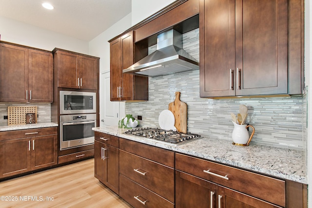 kitchen with light wood finished floors, appliances with stainless steel finishes, wall chimney exhaust hood, and light stone counters