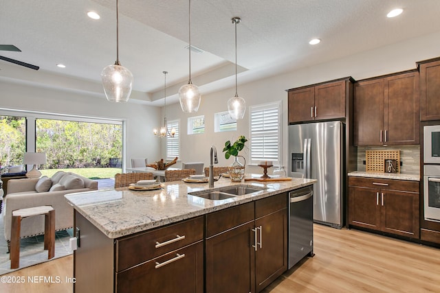 kitchen featuring a center island with sink, light wood finished floors, a tray ceiling, stainless steel appliances, and a sink