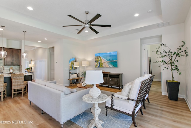 living area with light wood-style floors, visible vents, a tray ceiling, and a ceiling fan