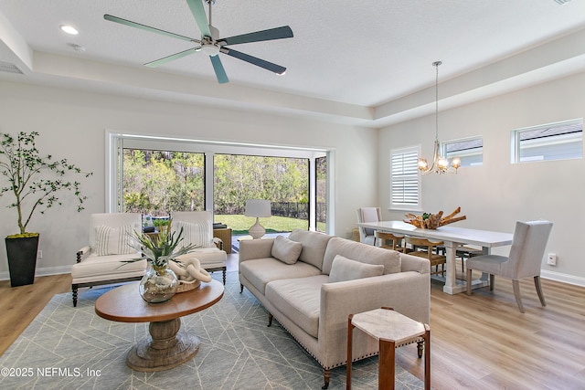 living room featuring a tray ceiling, ceiling fan with notable chandelier, baseboards, and light wood finished floors