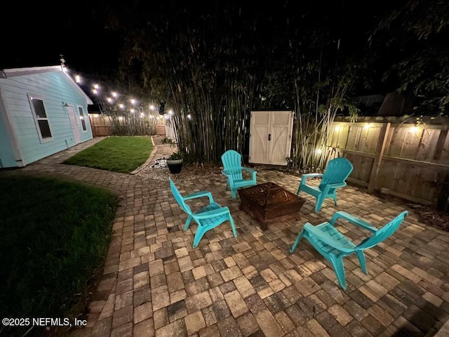 patio at night featuring an outdoor fire pit, a yard, a fenced backyard, an outdoor structure, and a storage unit