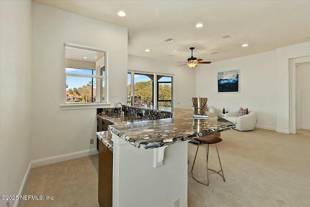 kitchen featuring visible vents, ceiling fan, light colored carpet, a breakfast bar, and dark stone countertops