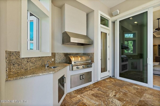 kitchen with a sink, light stone counters, stone finish flooring, wall chimney exhaust hood, and decorative backsplash