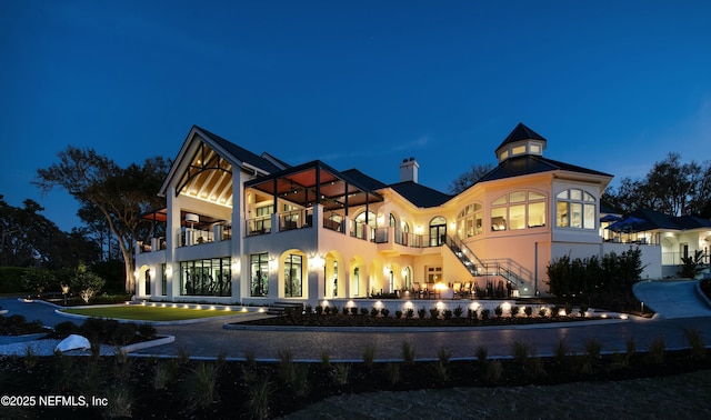 back of house at twilight with stucco siding, a balcony, and a chimney