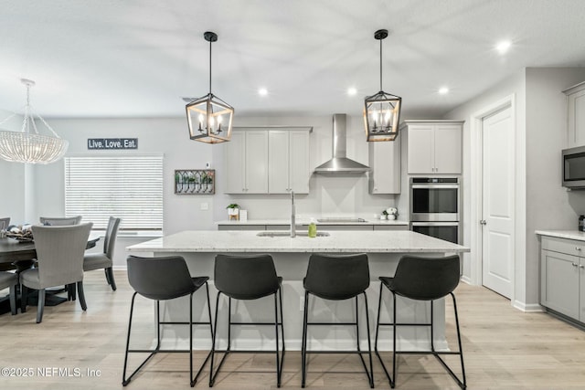 kitchen featuring light wood finished floors, wall chimney range hood, a chandelier, stainless steel appliances, and a sink