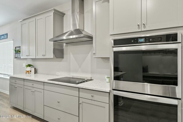 kitchen featuring gray cabinetry, double oven, wall chimney exhaust hood, light wood finished floors, and black electric cooktop