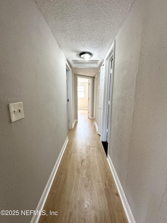 hall featuring light wood-style flooring, a textured wall, baseboards, and a textured ceiling