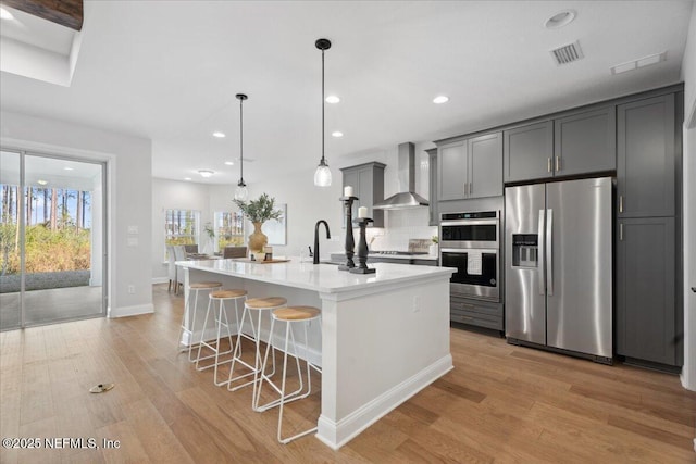 kitchen with light wood-type flooring, a center island with sink, a kitchen breakfast bar, appliances with stainless steel finishes, and wall chimney range hood