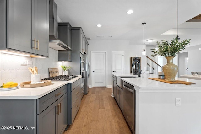 kitchen with light wood-style flooring, gray cabinets, a sink, stainless steel appliances, and wall chimney range hood