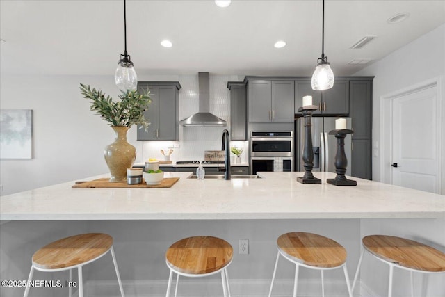 kitchen featuring gray cabinetry, a sink, appliances with stainless steel finishes, wall chimney exhaust hood, and a large island with sink