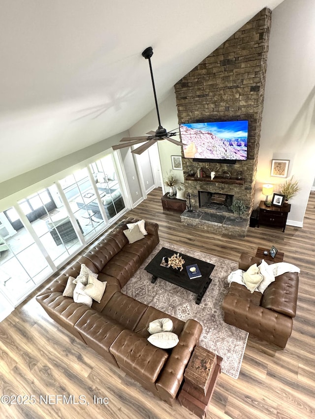 living area featuring high vaulted ceiling, a ceiling fan, wood finished floors, a stone fireplace, and baseboards