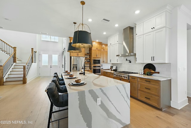 kitchen featuring light wood-style flooring, light stone counters, appliances with stainless steel finishes, wall chimney range hood, and decorative backsplash